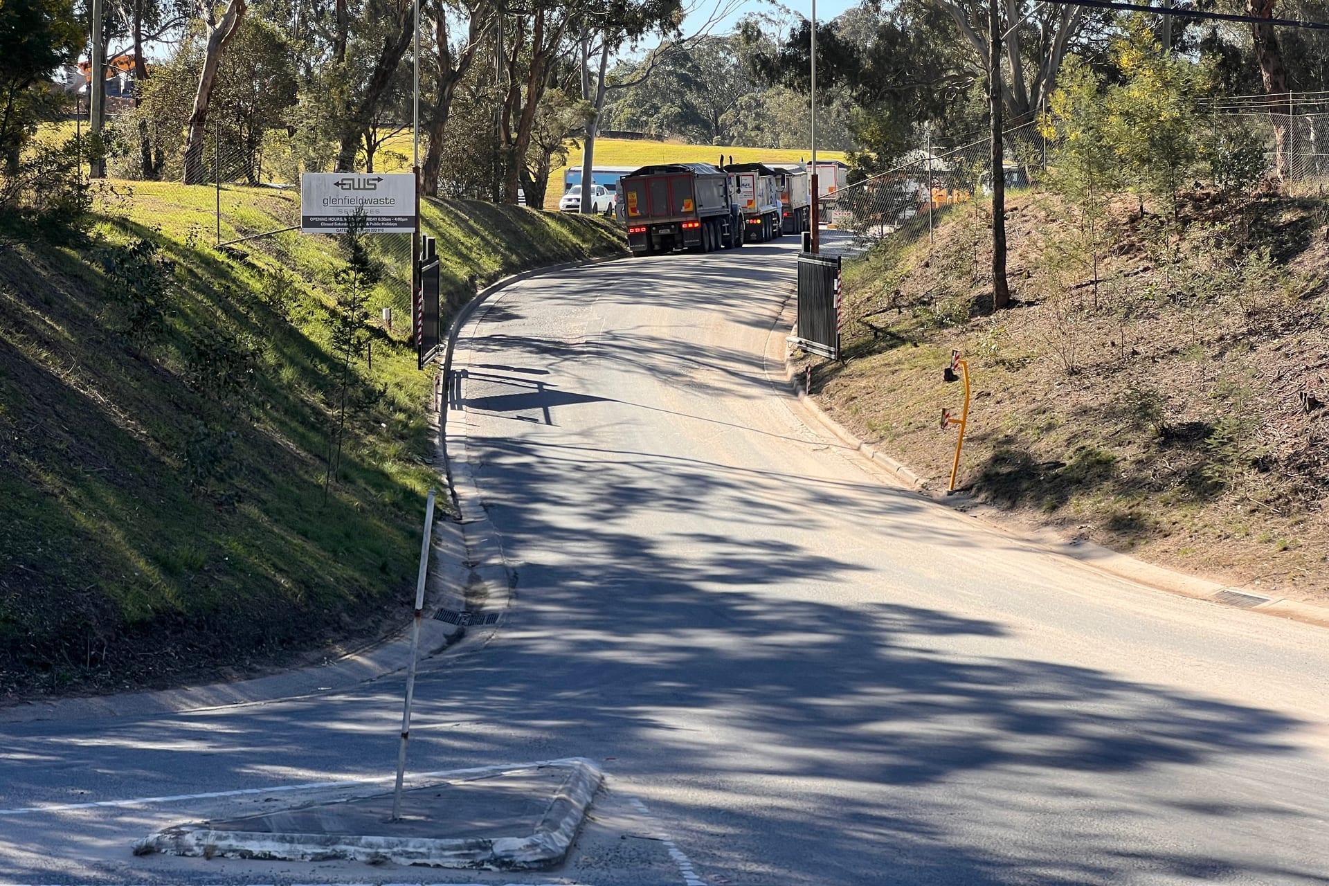The approach road into Glenfield Waste Services, with the site sign on the bank and trucks waiting at the boom gate.